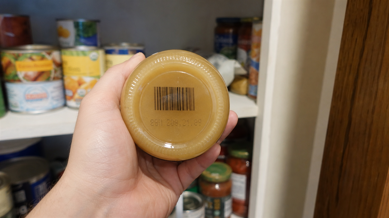 Person checking the best-by date and UPC code on an apple sauce jar in a home pantry during the Wholesome Pantry recall