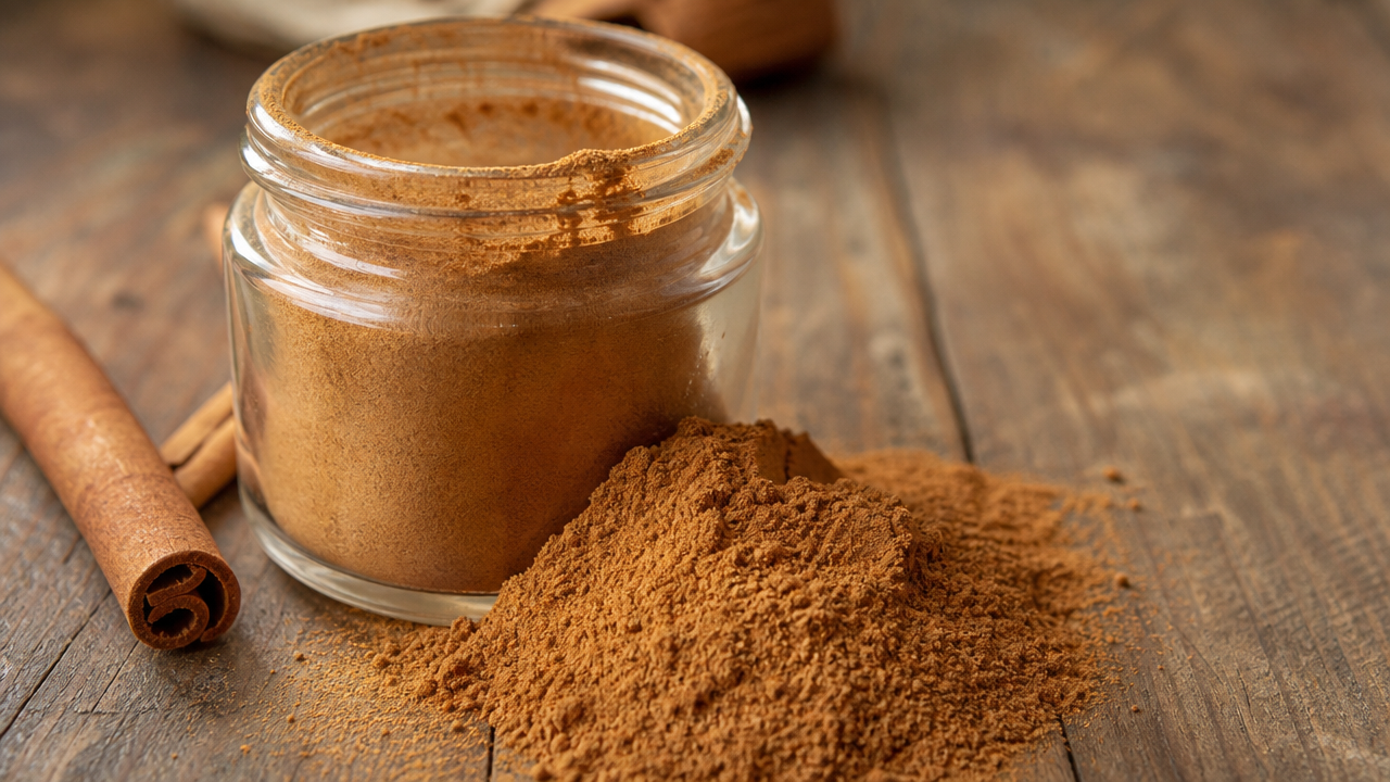 Ground cinnamon powder in a small glass jar next to a cinnamon stick on a wooden kitchen counter